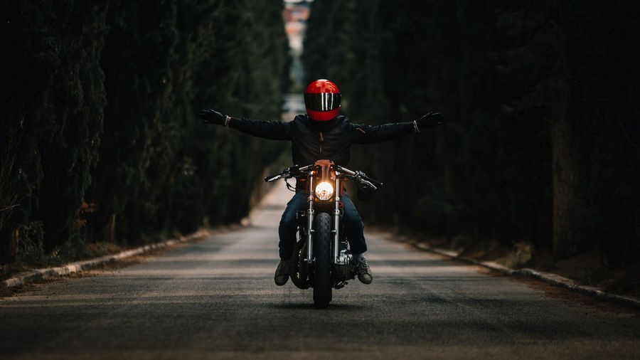 Biker in black leather jackets and helmet with open arms riding powerful motorcycle on asphalt road leading between green forest in countryside