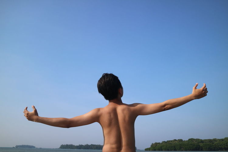 Rear view of shirtless man with arms outstretched against clear blue sky