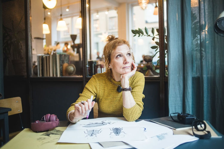 Thoughtful mature female illustrator looking away while sitting with drawing papers at desk in creative office