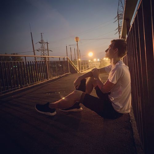 Side view of man sitting on bridge against sky during dusk