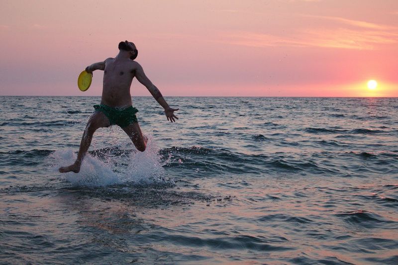 Playful shirtless man jumping in sea against orange sky