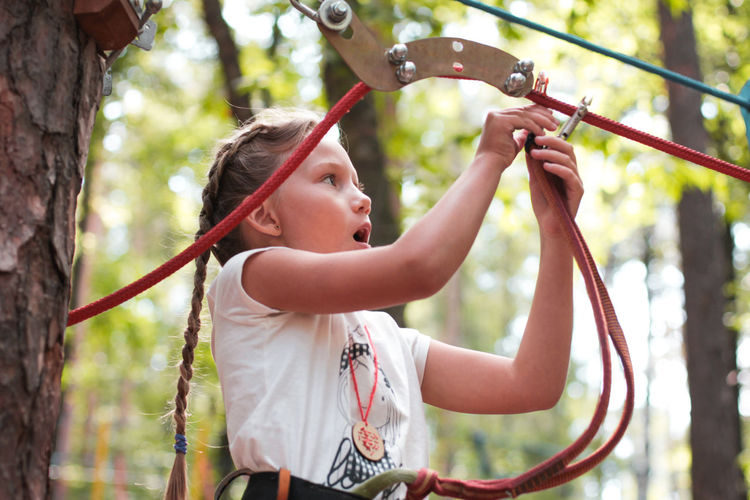 Low angle view of girl attaching safety harness on rope in forest