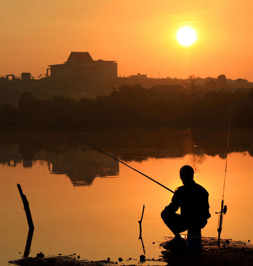 Silhouette man fishing by lake against orange sky during sunset