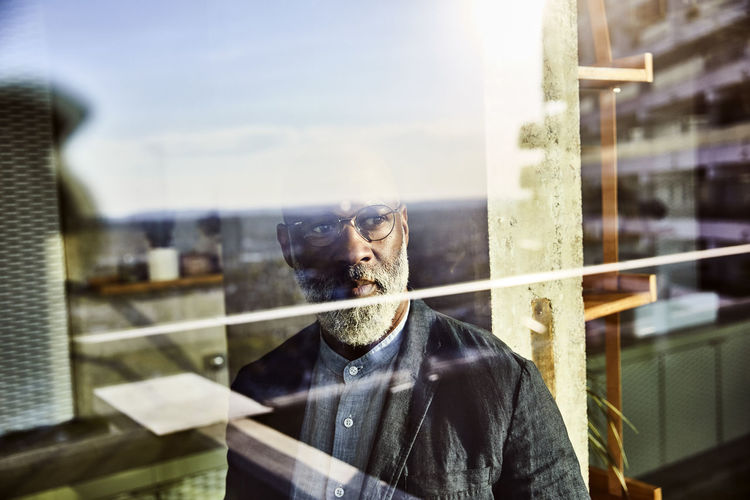 Reflection of mature mature man looking out of window while standing at home