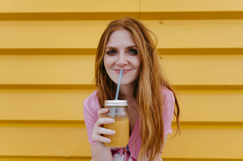 Smiling woman holding smoothie in front of yellow wall