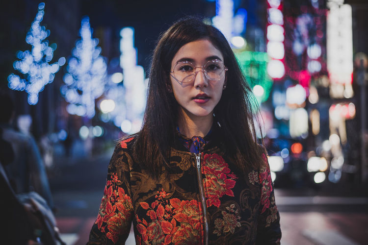 Portrait of young woman standing against illuminated city at night