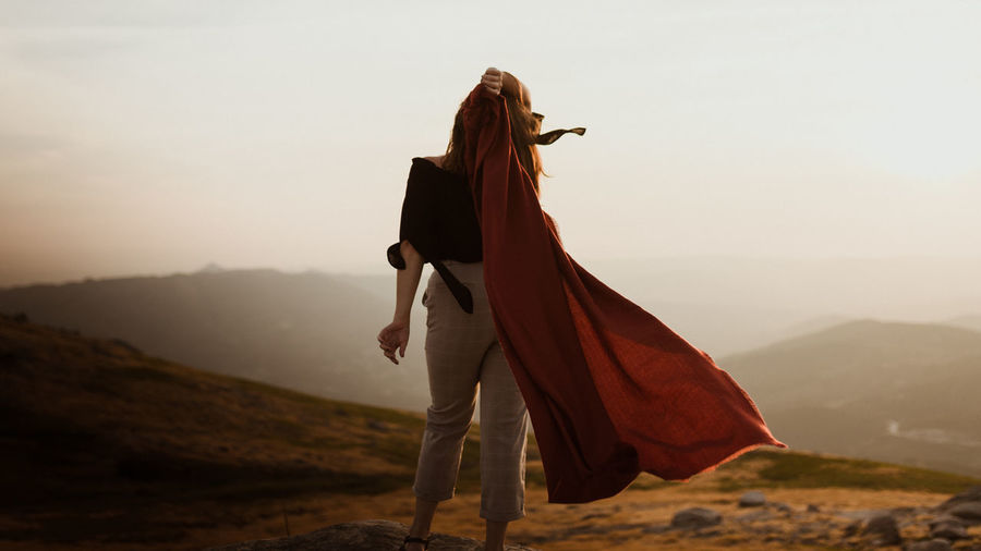 Woman standing on mountain against sky holding cape in wind