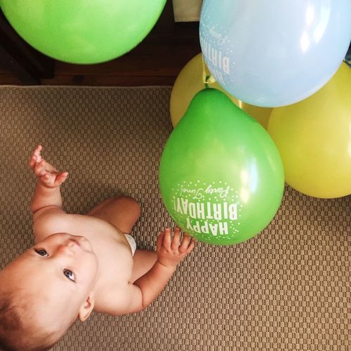 High angle view of shirtless baby boy kneeling by colorful balloons on carpet at home