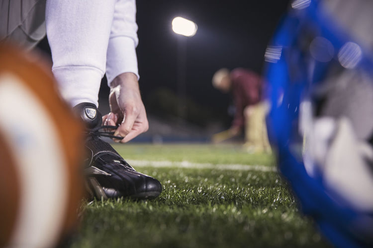 Low section of american football player tying shoelace on field with coach in background