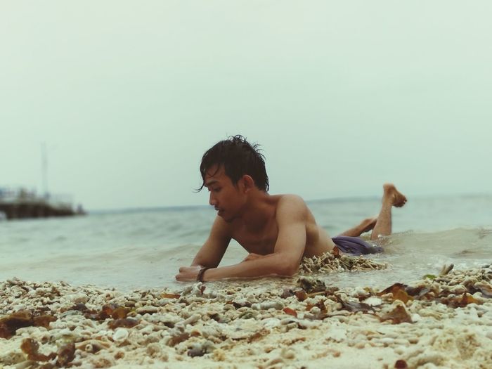 Shirtless young man lying at beach against sky