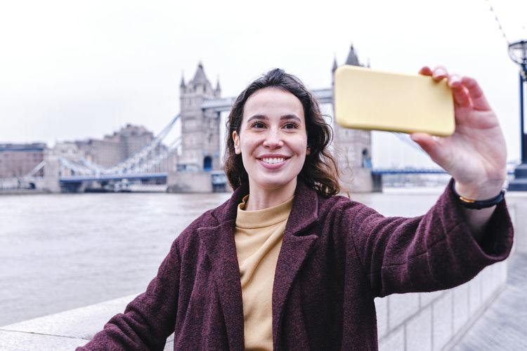 Smiling young woman taking selfie with tower bridge through smart phone