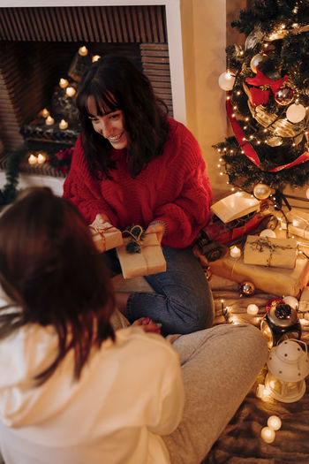 Smiling woman holding gifts while sitting with female friend at home during christmas