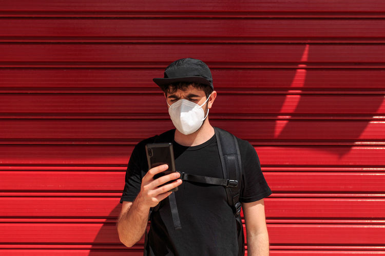 Young male in respiratory mask on cellphone near red wall standing on street during covid 19 pandemic