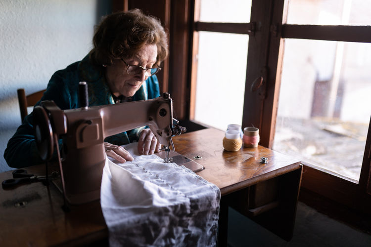 Senior female sewing white textile on special machine at table with spools of thread in room