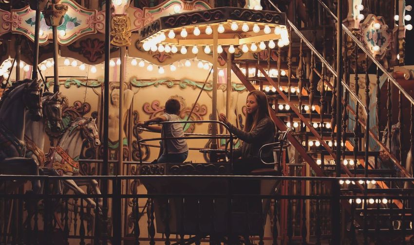 Siblings sitting in illuminated carousel horses at night