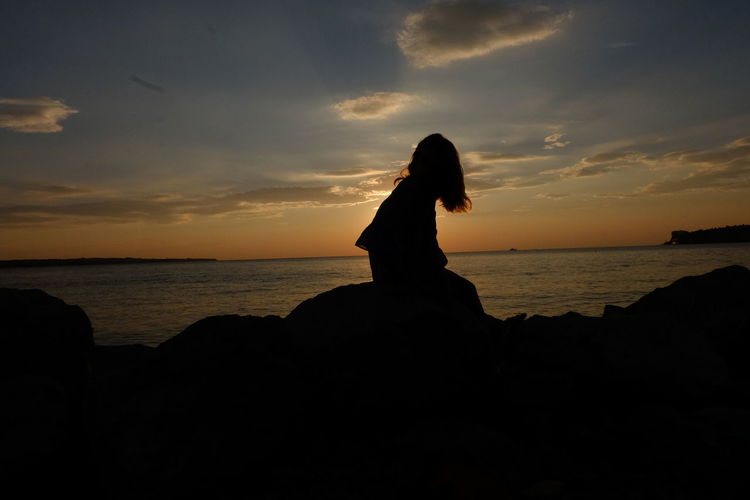 Silhouette teenage girls sitting on rock at beach against sky during sunset