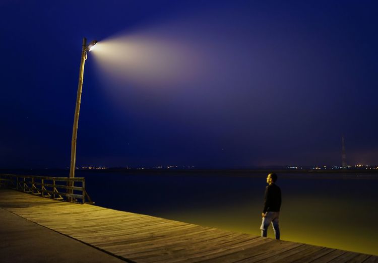 Rear view of man standing on illuminated pier over sea at night