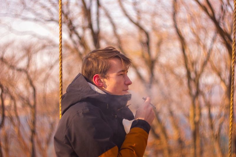 Side view of young man smoking cigarette against trees