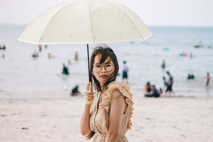 Portrait of beautiful woman wearing sunglasses holding umbrella standing at beach