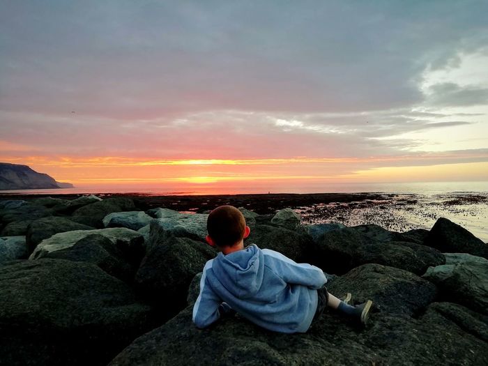 Rear view of boy sitting on rock by sea against sky