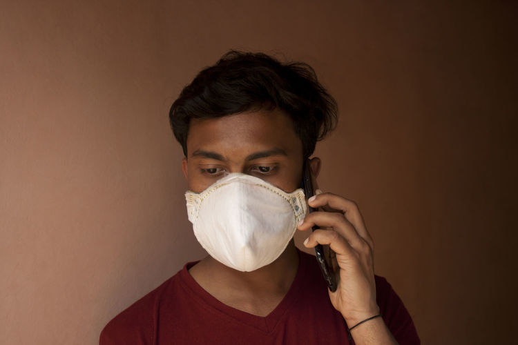 Close-up of young man wearing flu mask talking on phone against wall at home