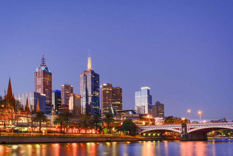 Illuminated city by princes bridge over yarra river against clear sky