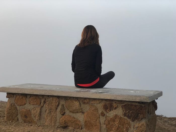 Rear view of woman sitting on retaining wall against sky