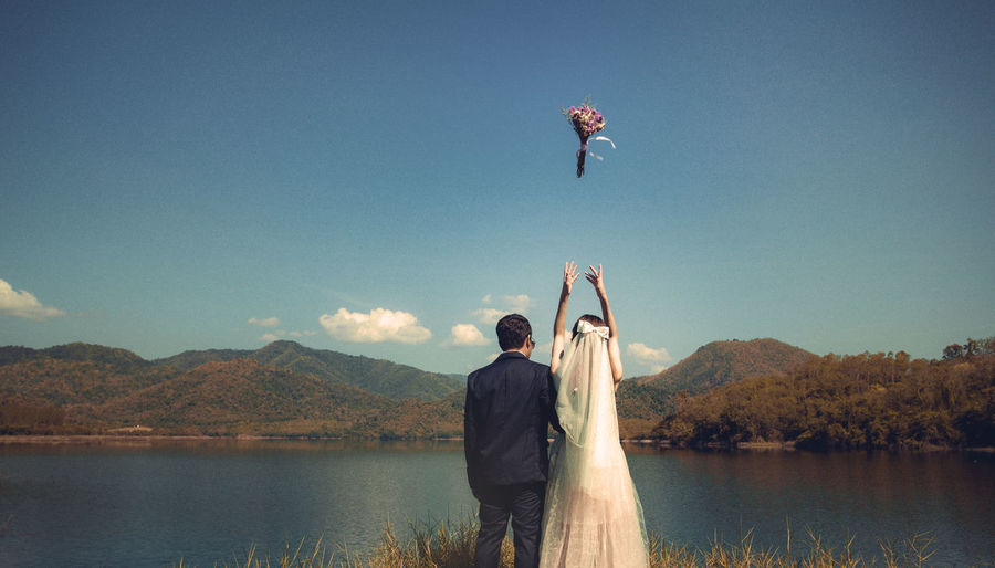Couple throwing bouquet while standing at lakeshore against sky