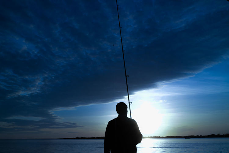 Silhouette man fishing in sea against cloudy sky