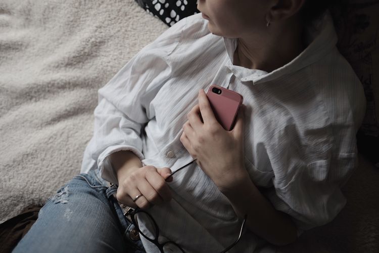 Midsection of woman holding pink mobile phone resting on bed at home