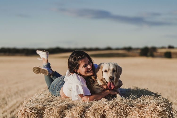 Happy mid adult woman with dog lying on hay at agricultural field
