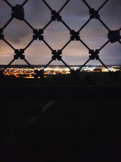 Illuminated cityscape seen through chainlink fence