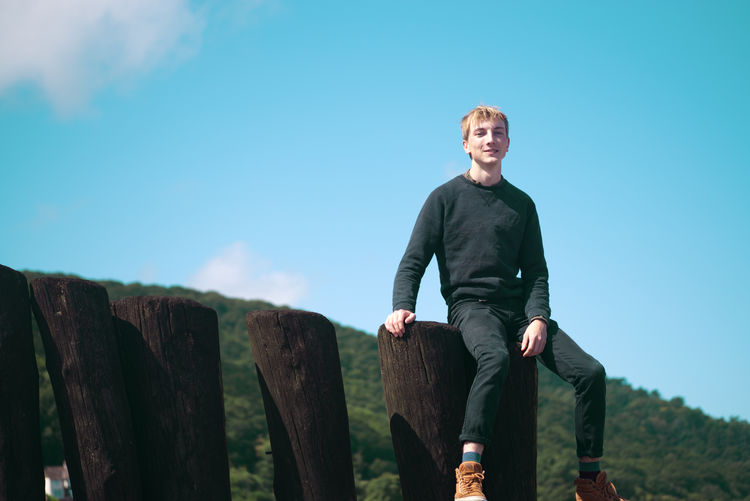 Portrait of young man sitting on wooden logs against sky