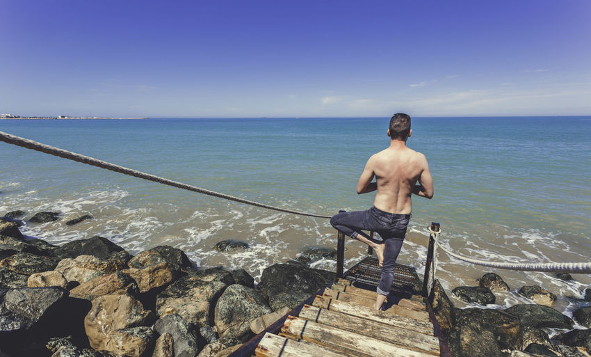 Rear view of shirtless man doing yoga on steps by sea against clear blue sky