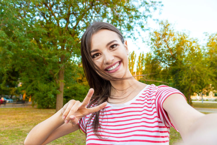 Portrait of smiling beautiful woman showing peace sign at park