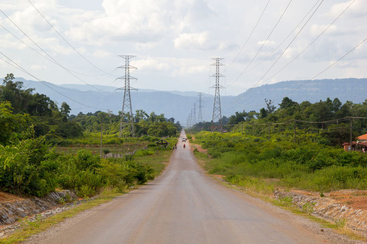 Road amidst trees and electricity pylons against sky
