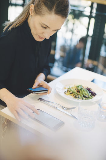 Confident female entrepreneur using mobile phone while sitting with credit card at cafeteria in office