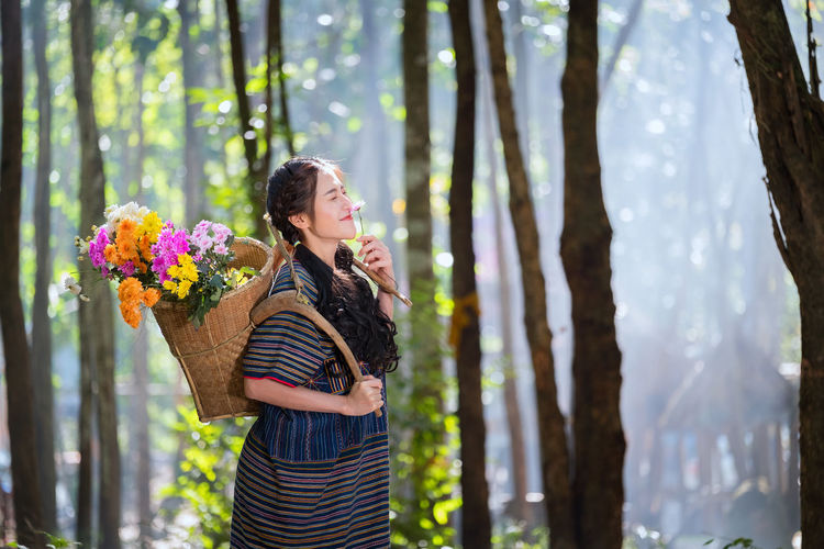 Smiling beautiful young woman carrying flowers basket while standing in forest