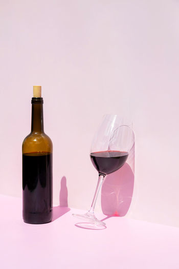 A bottle of red wine and a filled glass next to it in the sun on a pink background. place for text