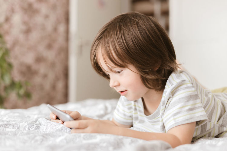 A cute boy, toddler, lies on the bed in the bedroom and plays with the phone, smartphone. 
