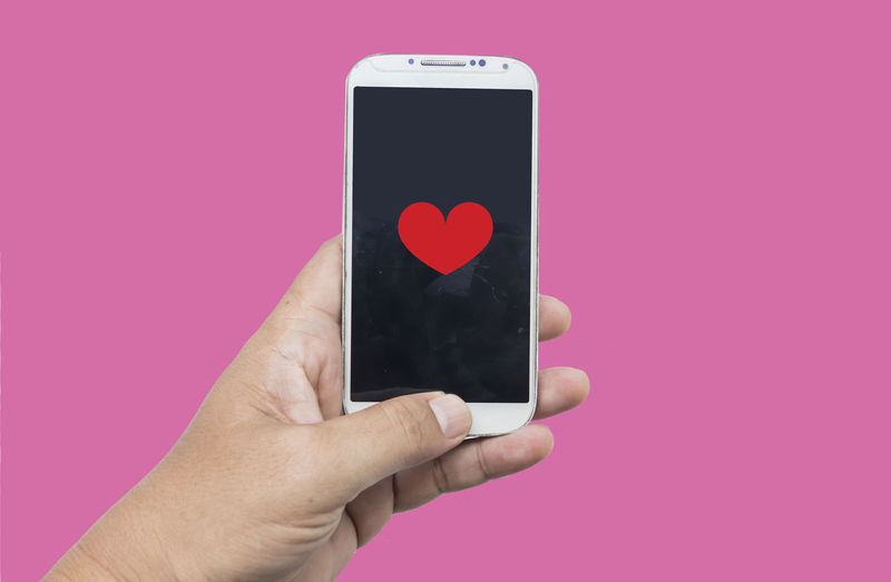 Close-up of hand holding smart phone against pink background
