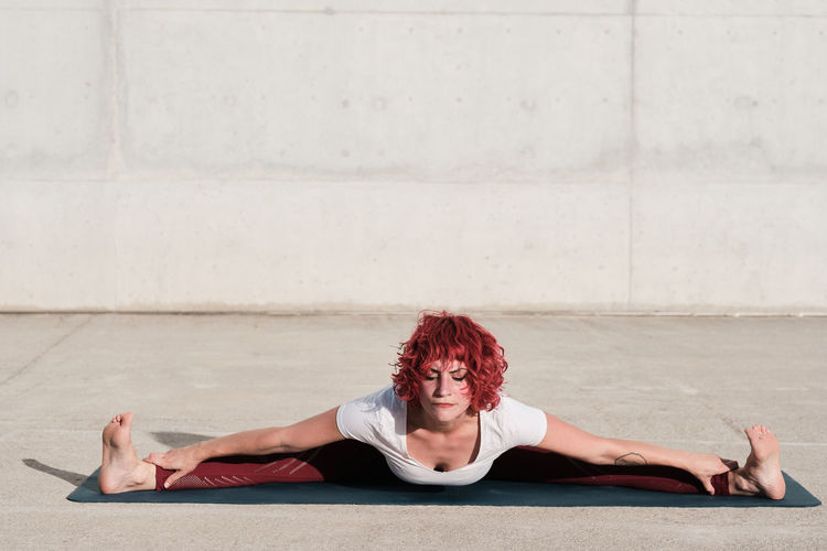 From above of barefooted woman with with closed eyes in sportswear doing yoga in wide angle seated forward bend pose on mat training alone on street against concrete wall