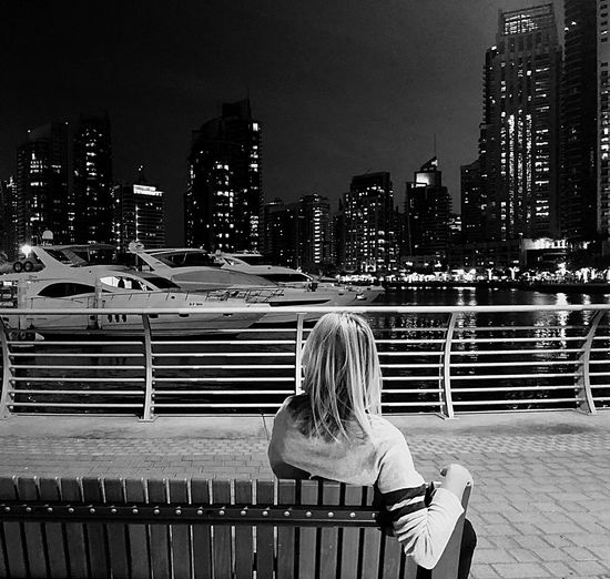 Rear view of woman sitting on bench against illuminated buildings in city at night