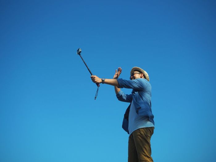Low angle view of happy man taking selfie with monopod against clear blue sky
