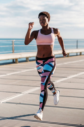 From below african american adult sportswoman in vibrant green activewear focusing and running alone along waterfront among metal columns under roof