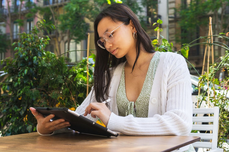 Charming asian female freelancer writing on tablet with stylus while sitting on terrace of cafeteria on summer day in city