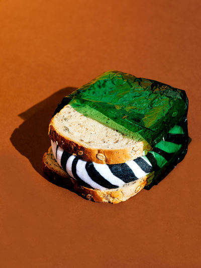 Sandwich in green harmful environmentally unfriendly wrap and with zebra filling placed on brown surface near brown wall in light studio