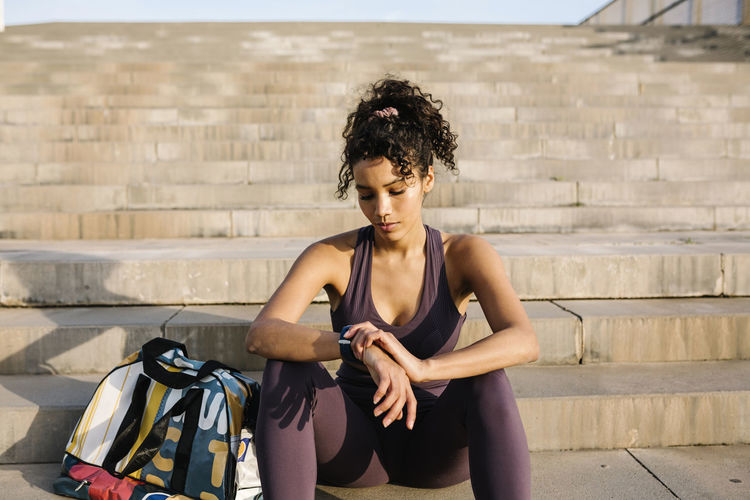 Female athlete checking time while sitting by bag on staircase on sunny day