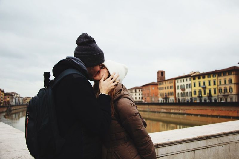 Couple kissing while standing by canal against sky in city