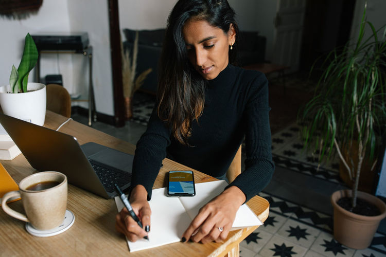High angle of indian female entrepreneur sitting at table and writing plans in notebook while working at home in cozy workplace